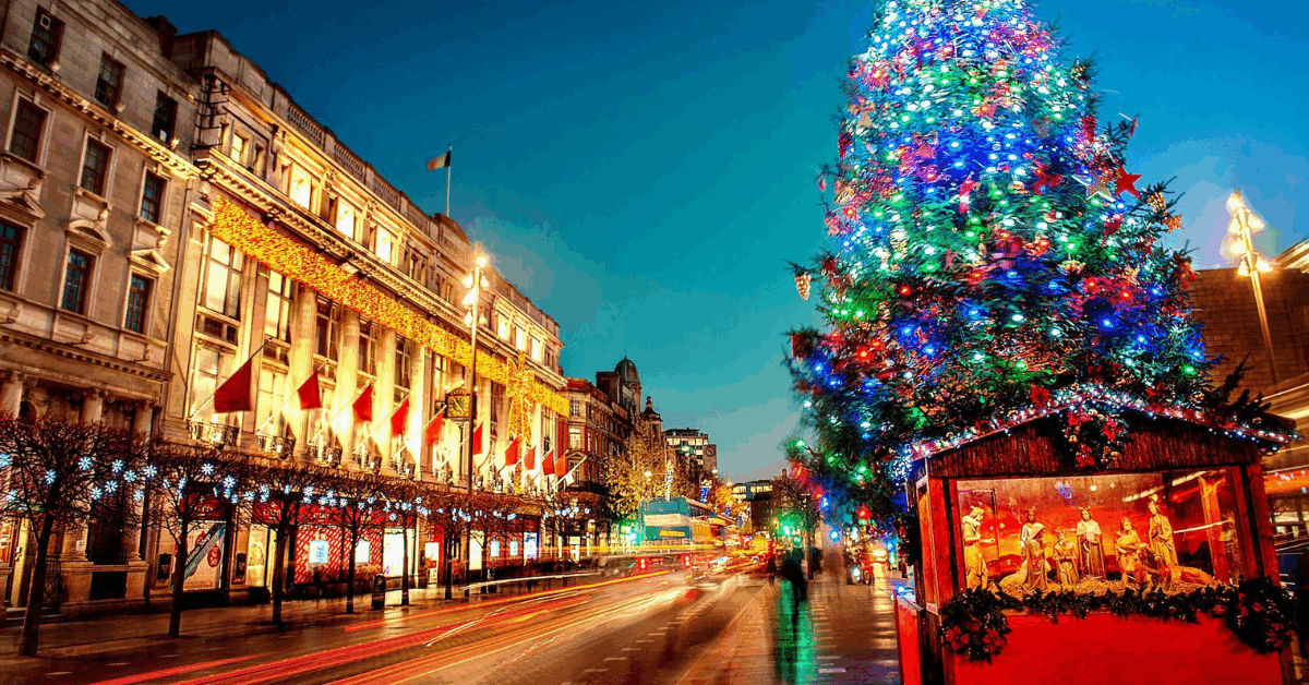 A brightly lit Christmas tree stands beside a busy street lined with decorated buildings and holiday lights at dusk.
