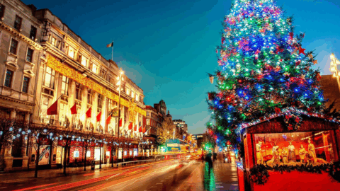 A brightly lit Christmas tree stands beside a busy street lined with decorated buildings and holiday lights at dusk.