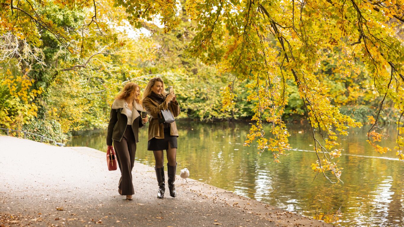 Two women walk together along a path beside a lake in a park with autumn foliage, holding coffee cups and bags, under overhanging yellow and green tree branches.
