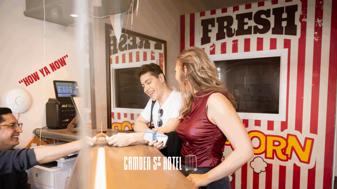 Two people check in at a hotel reception with a popcorn-themed backdrop while a receptionist hands them a room key—perfect for those seeking fun things to do in summer. The words